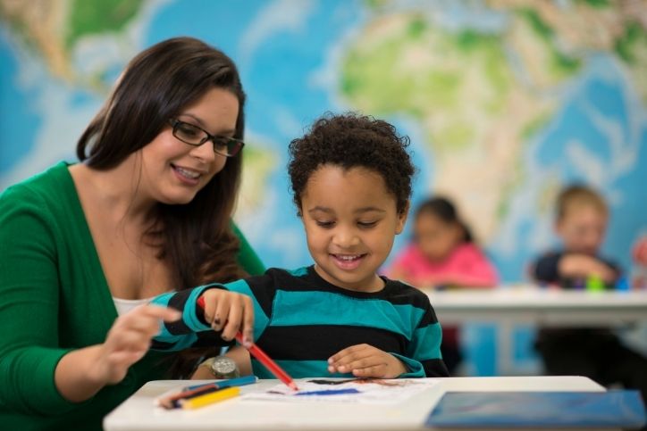 Uma professora sorridente está ao lado de um menino pequeno, que está sentado em uma mesa de aula. Ambos estão olhando e interagindo com um desenho que o menino está colorindo. Ao fundo, um mapa-múndi azul decora a parede, e outras crianças estão desfocadas ao fundo.