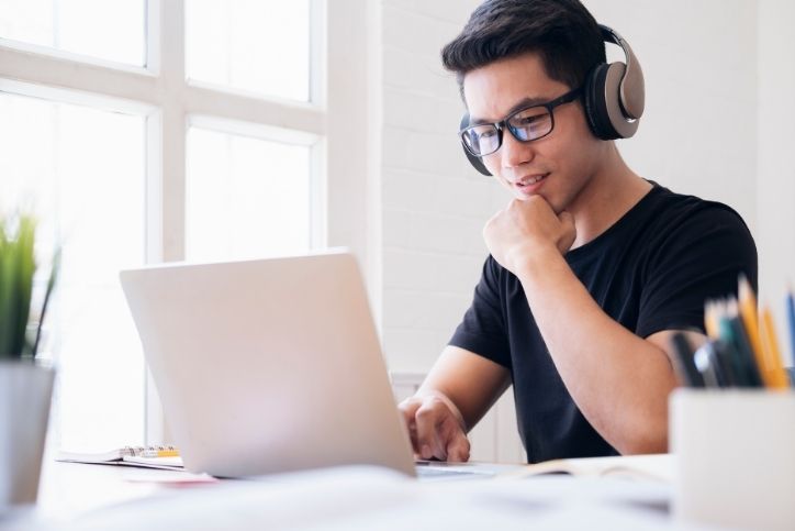 Um jovem usando fones de ouvido e óculos, sentado em frente ao laptop, em uma mesa com luz natural ao fundo.